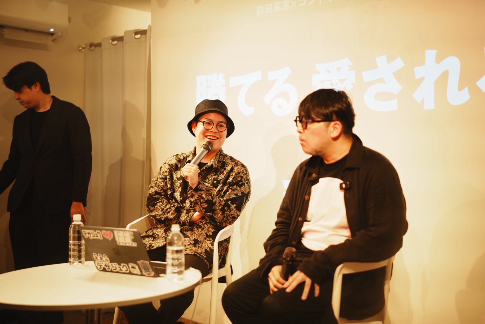 「勝てる 愛される」のスクリーンを背景に語り合う前田高志氏とコンドウハルキ